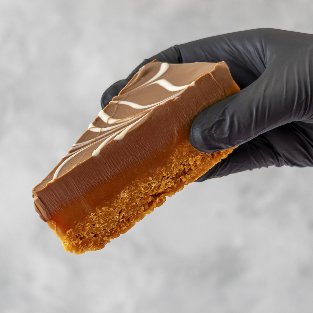 Millionaire Cornflake Pie Slice held in black glove with smooth chocolate topping