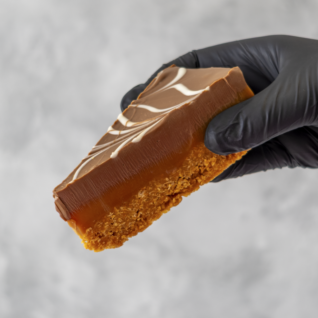 Millionaire Cornflake Pie Slice held in black glove with caramel layer and white chocolate drizzle