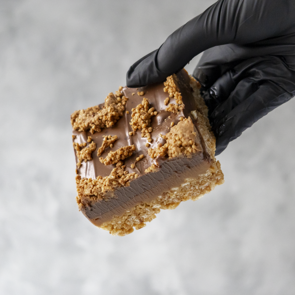Hand wearing a black glove holding a chocolate dessert with crumbly topping against a neutral background