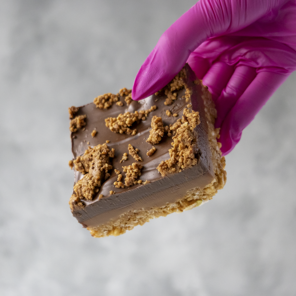 DoughGirl Cracking Caramel Crispie Slab with buttery caramel, golden crisped rice, and silky milk chocolate, held by a hand in a pink glove on a neutral background
