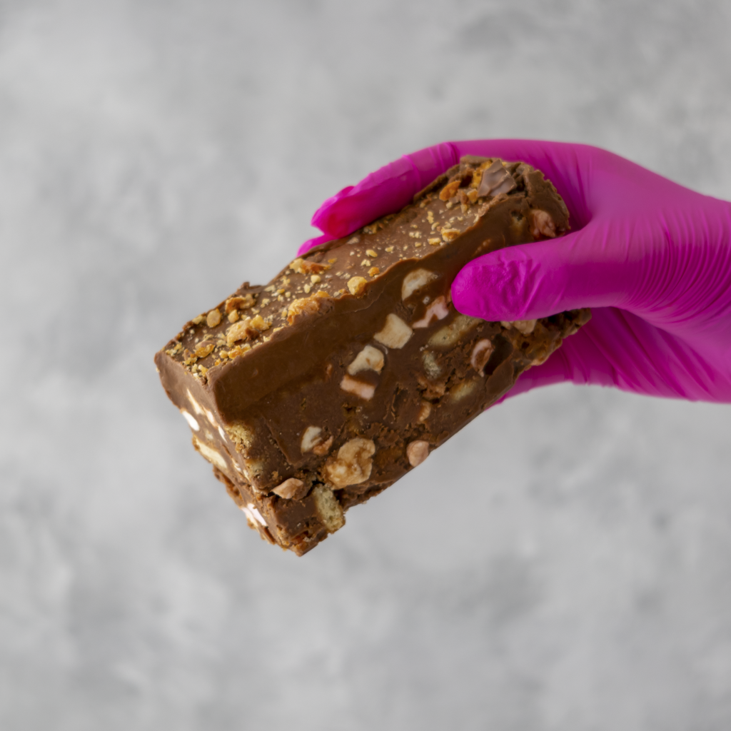 Hand wearing a pink glove holding a Crunchie Rocky Mountain Road cake against a gray background