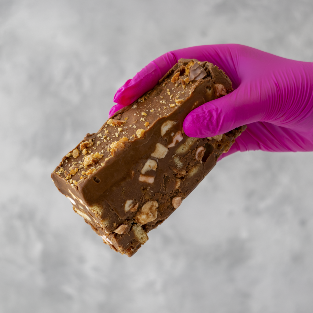 Hand wearing a purple glove holding a Crunchie Rocky Mountain Road cake against a gray background