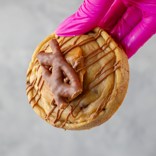Curly Wurly Deluxe Cookie with chocolate drizzle and a piece of chocolate on top, held by a hand in a pink glove.