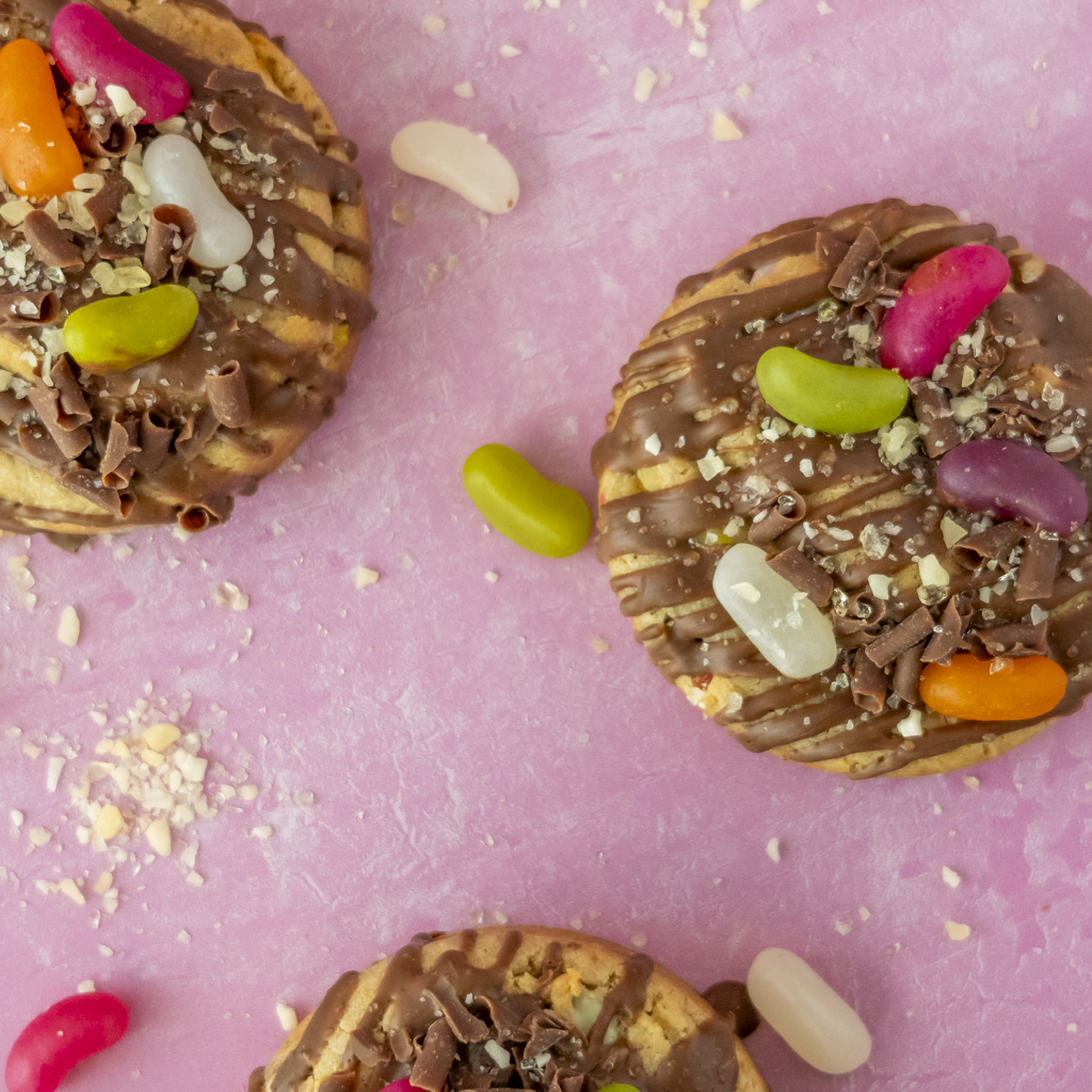 Three round chocolate-covered cookies topped with drizzled chocolate, crumbled chocolate, and colourful jelly beans, arranged on a pink background with scattered crumbs and loose jelly beans.