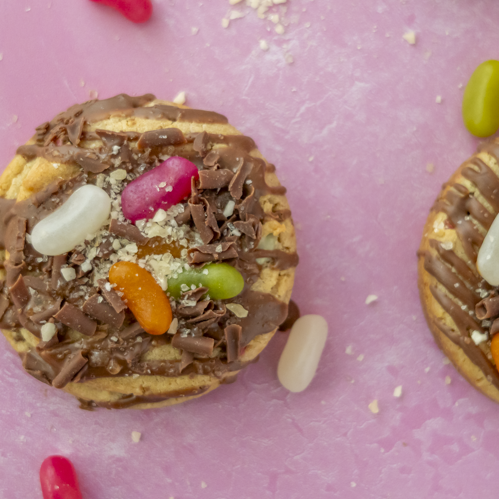 A close-up of a round chocolate-covered cookie topped with chocolate shavings, crushed crumbs, and colourful jelly beans, set on a pink surface with more beans and part of another cookie visible at the right edge.