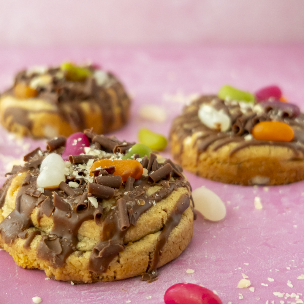 Jelly Pop Deluxe Cookies topped with milk chocolate drizzle, colourful jelly sweets, chocolate curls and white chocolate pieces on a pink background.