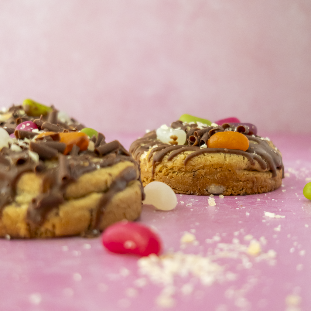Two chocolate-dipped cookies topped with colourful candy pieces, chocolate curls, and white sprinkles, arranged on a pink surface with a soft pink background.
