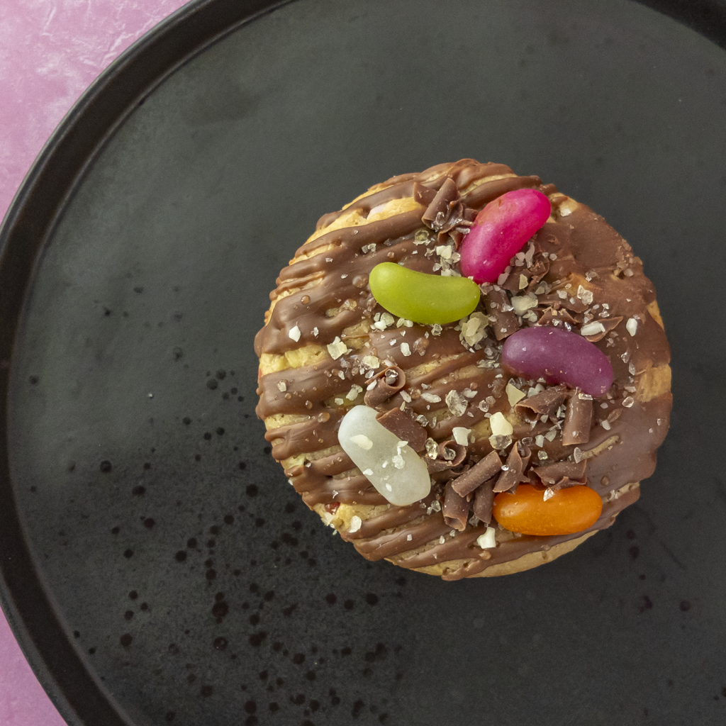 A round chocolate-covered cookie topped with drizzles of chocolate, chocolate shavings, coarse sugar crystals, and five colourful jelly beans, presented on a dark round plate against a pink background.