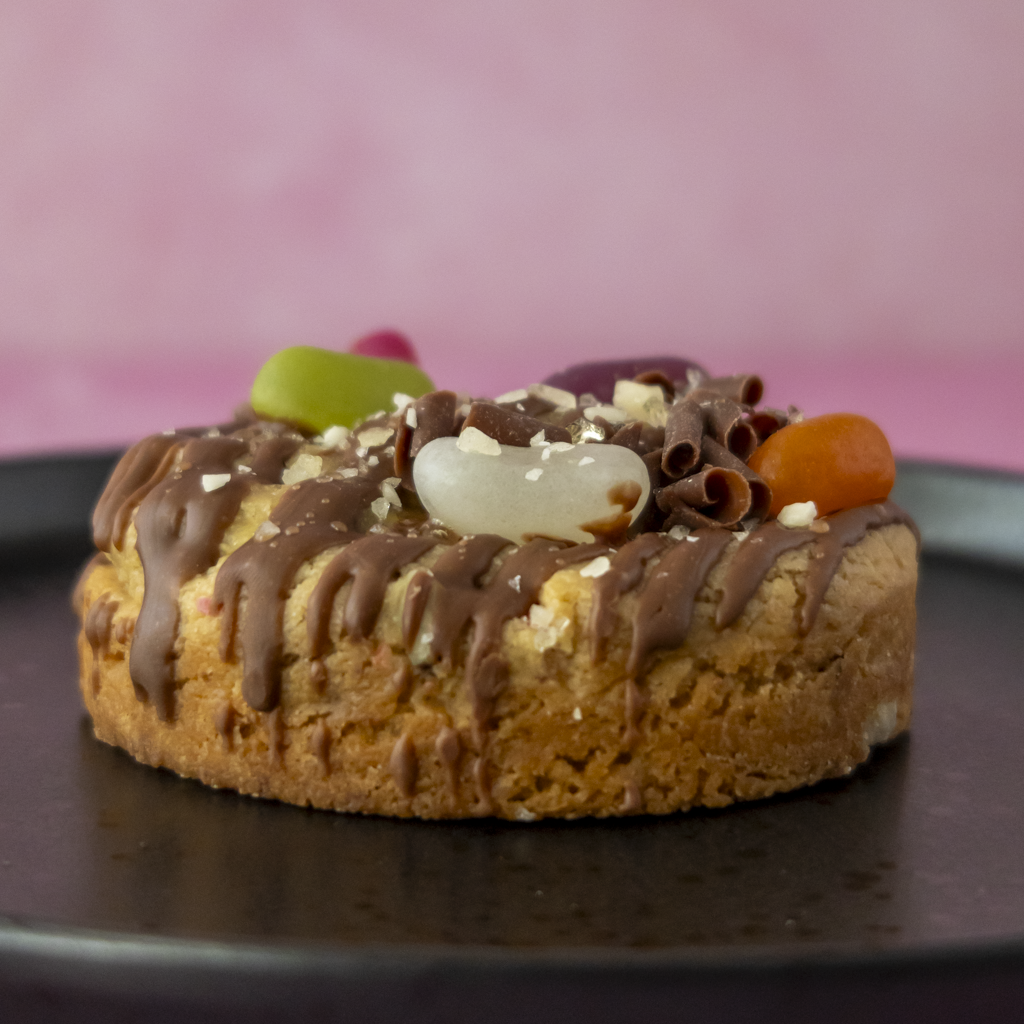 A small round chocolate cookie topped with dark chocolate drizzle, chocolate shavings, crushed nuts, and colourful candy pieces, presented on a dark plate against a pink background.