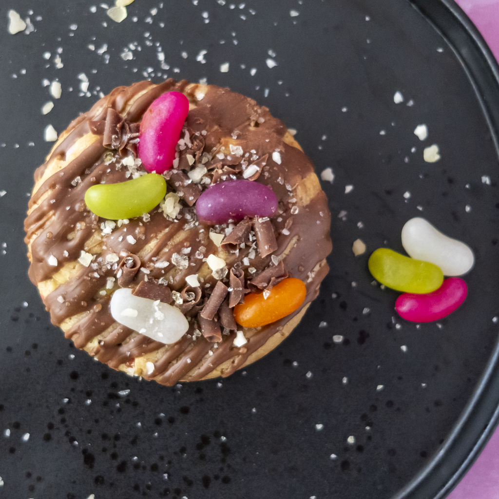A round chocolate-topped cookie on a black plate, decorated with drizzled chocolate, chocolate shavings, coarse flakes, and several colourful jelly beans.
