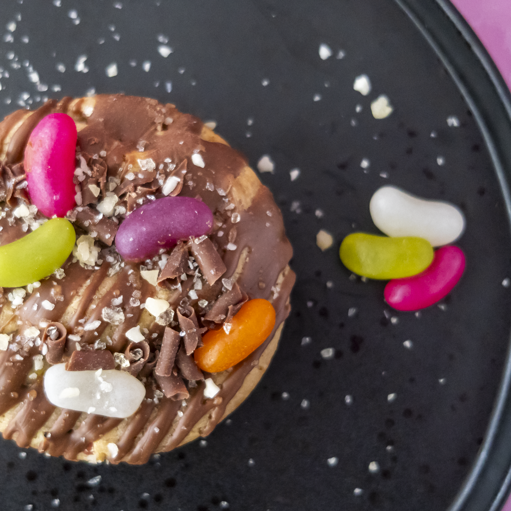A close-up overhead shot of a chocolate-covered cookie topped with chocolate shavings, coarse flakes, and several colourful jelly beans, set on a dark plate with a few loose jelly beans nearby.