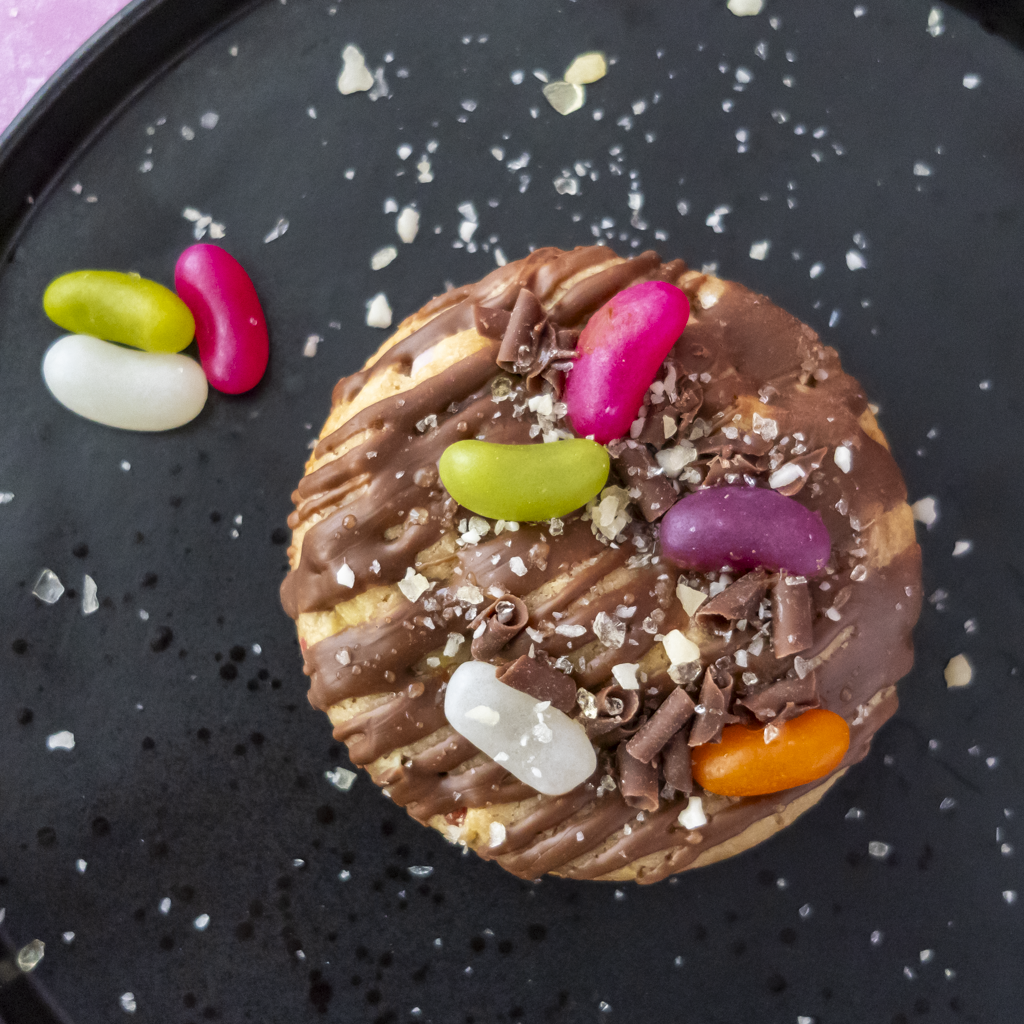 A round chocolate-covered cookie topped with drizzles of chocolate, crushed chocolate shavings, coarse salt flakes, and several colourful jelly beans, presented on a black plate.