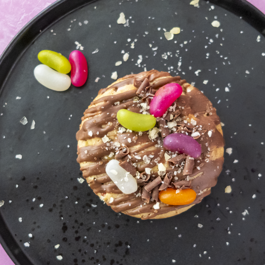 A round chocolate-covered cookie topped with drizzles, chocolate shavings, and colourful jelly beans, presented on a black plate with a few loose jelly beans nearby.