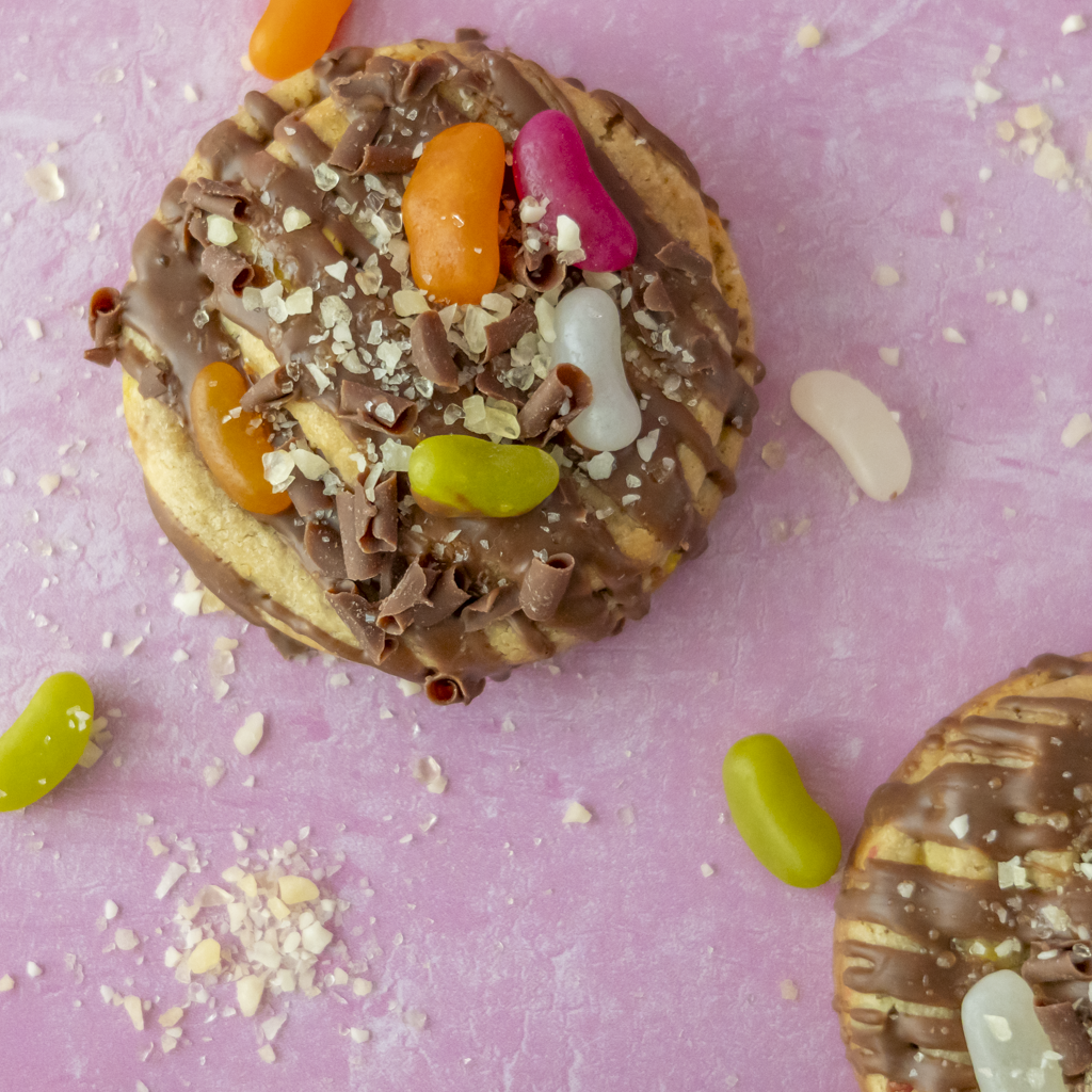 Two chocolate-covered cookies topped with crumbled chocolate, white chocolate shavings, and colourful jelly beans, arranged on a pink background with scattered crumbs and loose jelly beans.