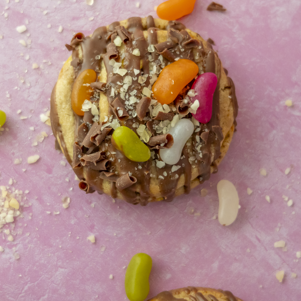 Jelly Pop Deluxe Cookie topped with milk chocolate drizzle, colourful jelly sweets, chocolate curls and white chocolate pieces on a pink background.