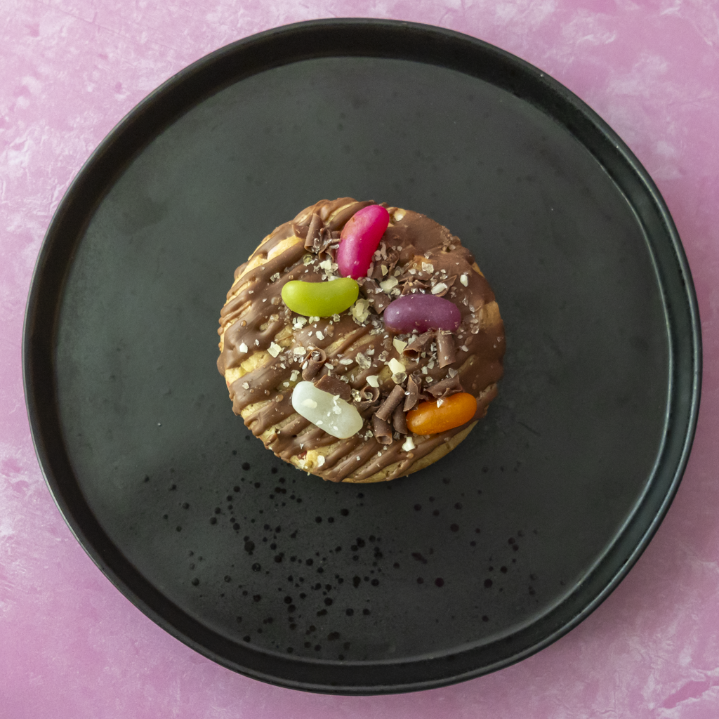 A round chocolate-covered cookie  topped with drizzles, chopped nuts, and colourful jelly beans, centred on a black plate against a pink background.