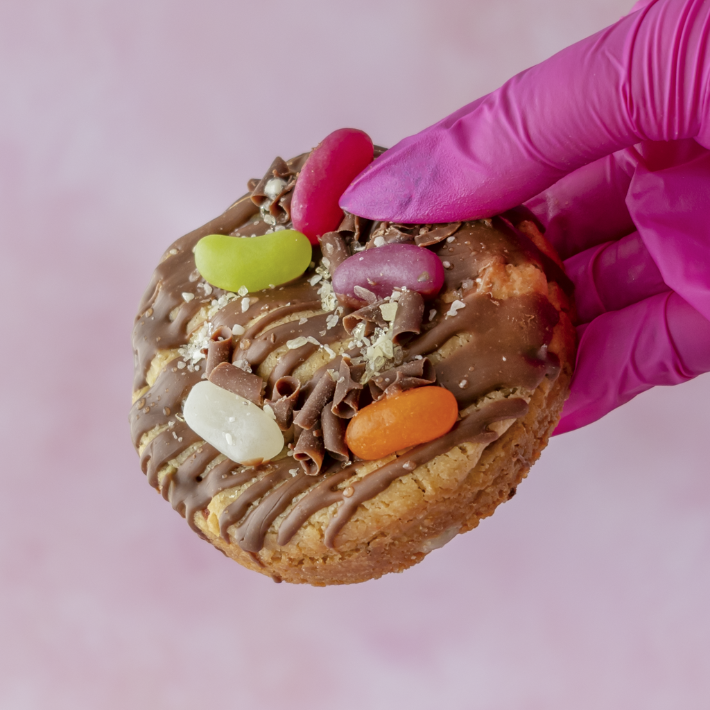 Jelly Pop Deluxe Cookie topped with milk chocolate drizzle, colourful jelly pops, chocolate curls and popping candy, held in a pink gloved hand.