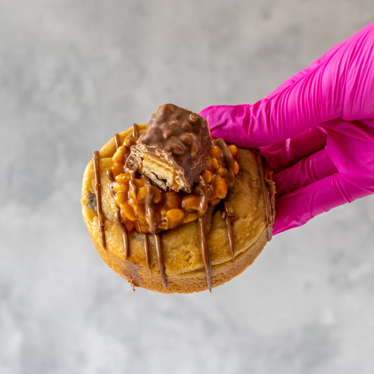 Lion Bar Stuffed Deluxe Cookie with chocolate drizzle and a piece of cookie held by a gloved hand against a neutral background