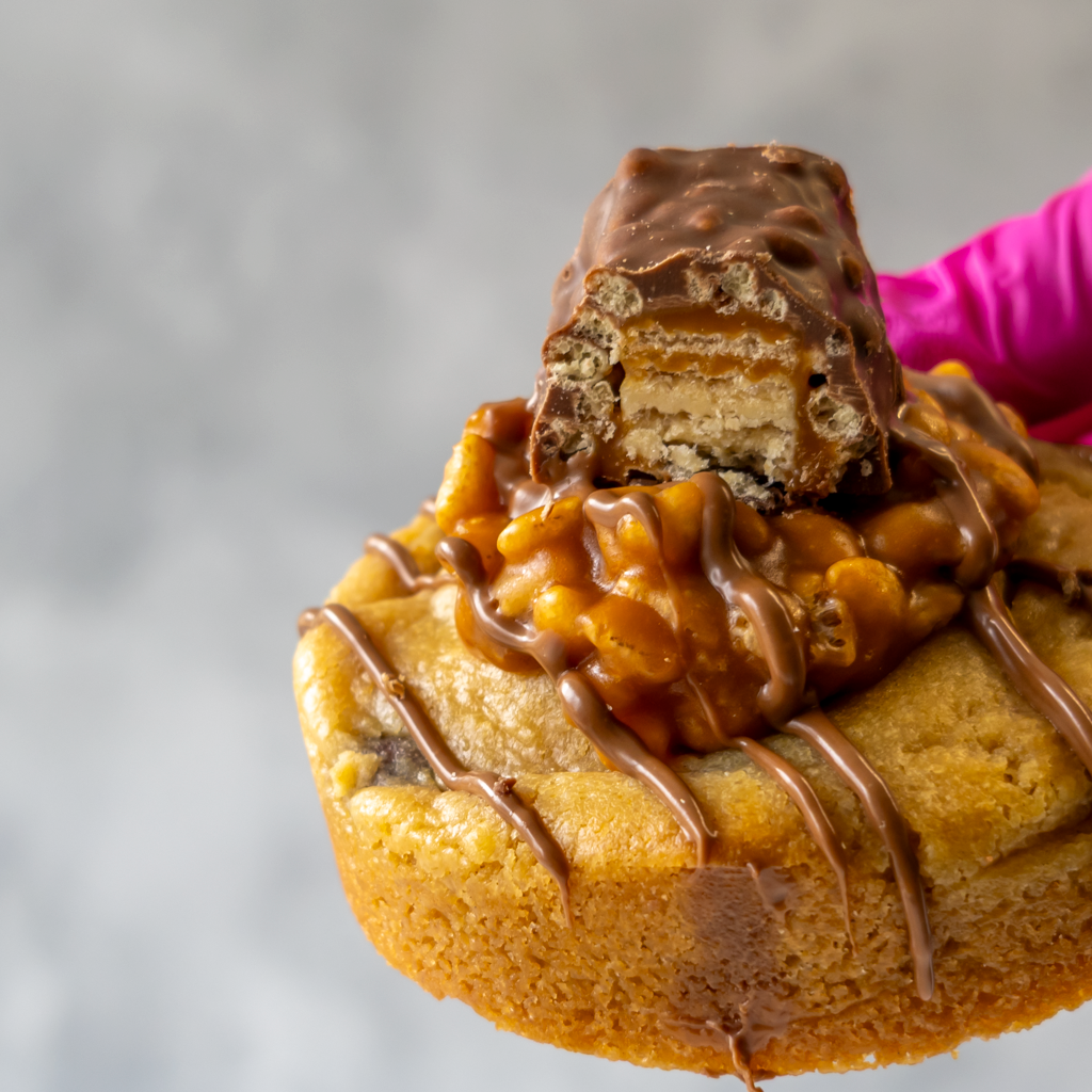 Close-up of a Lion Bar Stuffed Deluxe Cookie with chocolate drizzle and a Lion bar on top against a blurred background