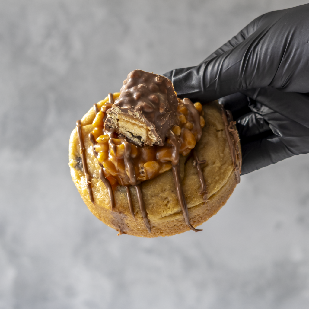 Lion Bar Stuffed Deluxe Cookie with chocolate drizzle and a piece of candy bar held by a gloved hand against a gray background