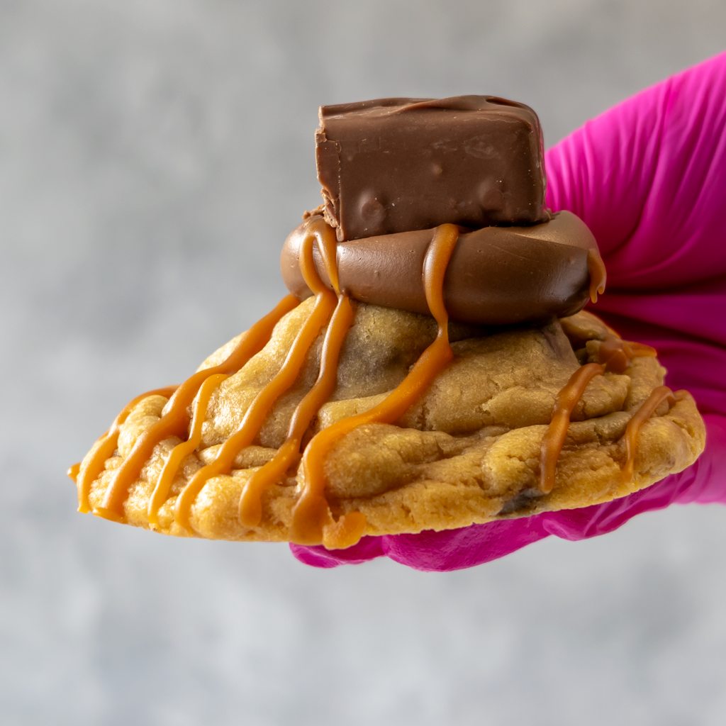 Mars Deluxe Cookie with caramel and chocolate drizzle held by a pink gloved hand against a gray background