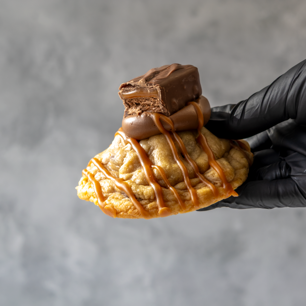Hand holding a Mars Deluxe Cookie with chocolate and caramel drizzle against a gray background