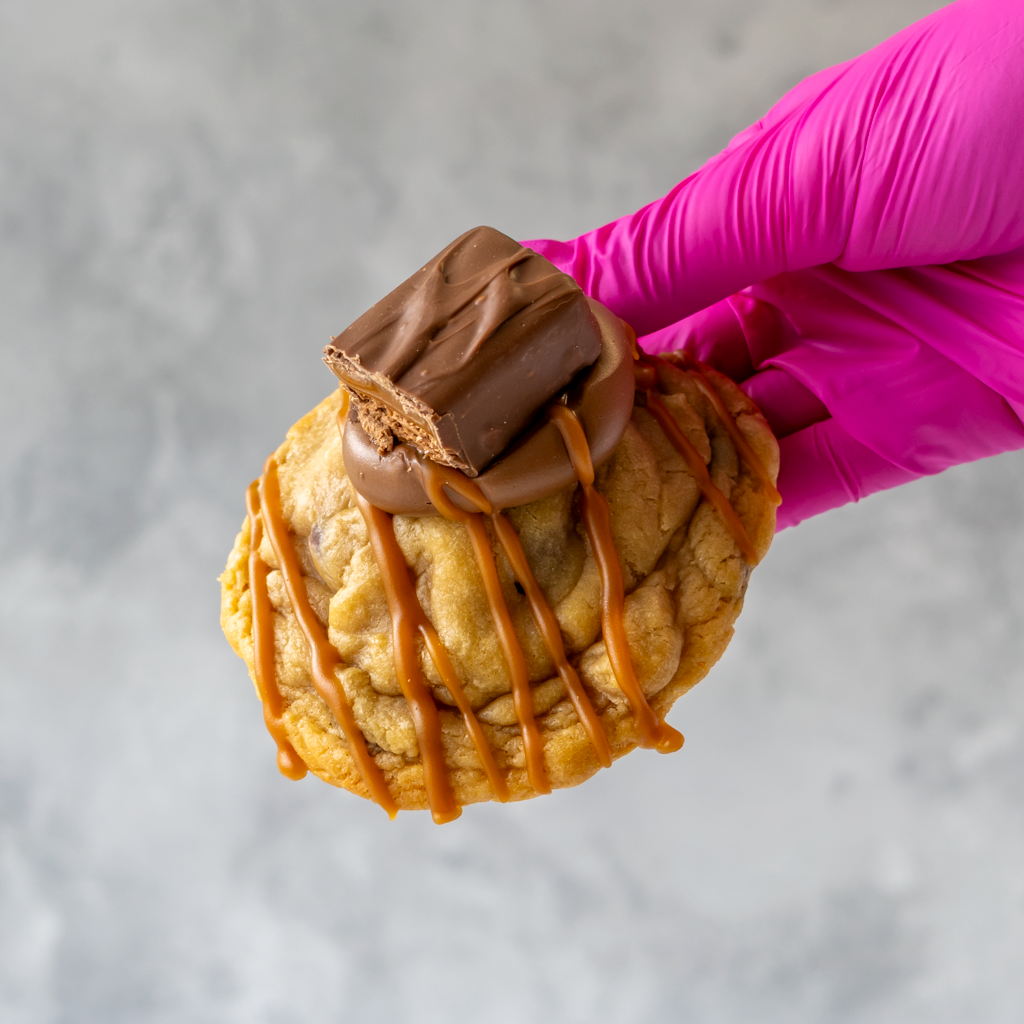 Hand holding a Mars Deluxe Cookie with caramel drizzle and chocolate bar against a neutral background