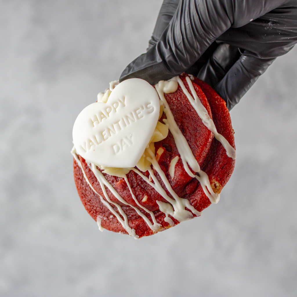 Limited Edition Valentine's Red Velvet Deluxe Cookie with white chocolate drizzle and a heart-shaped Valentine's Day topper, held by a black gloved hand against a gray background.