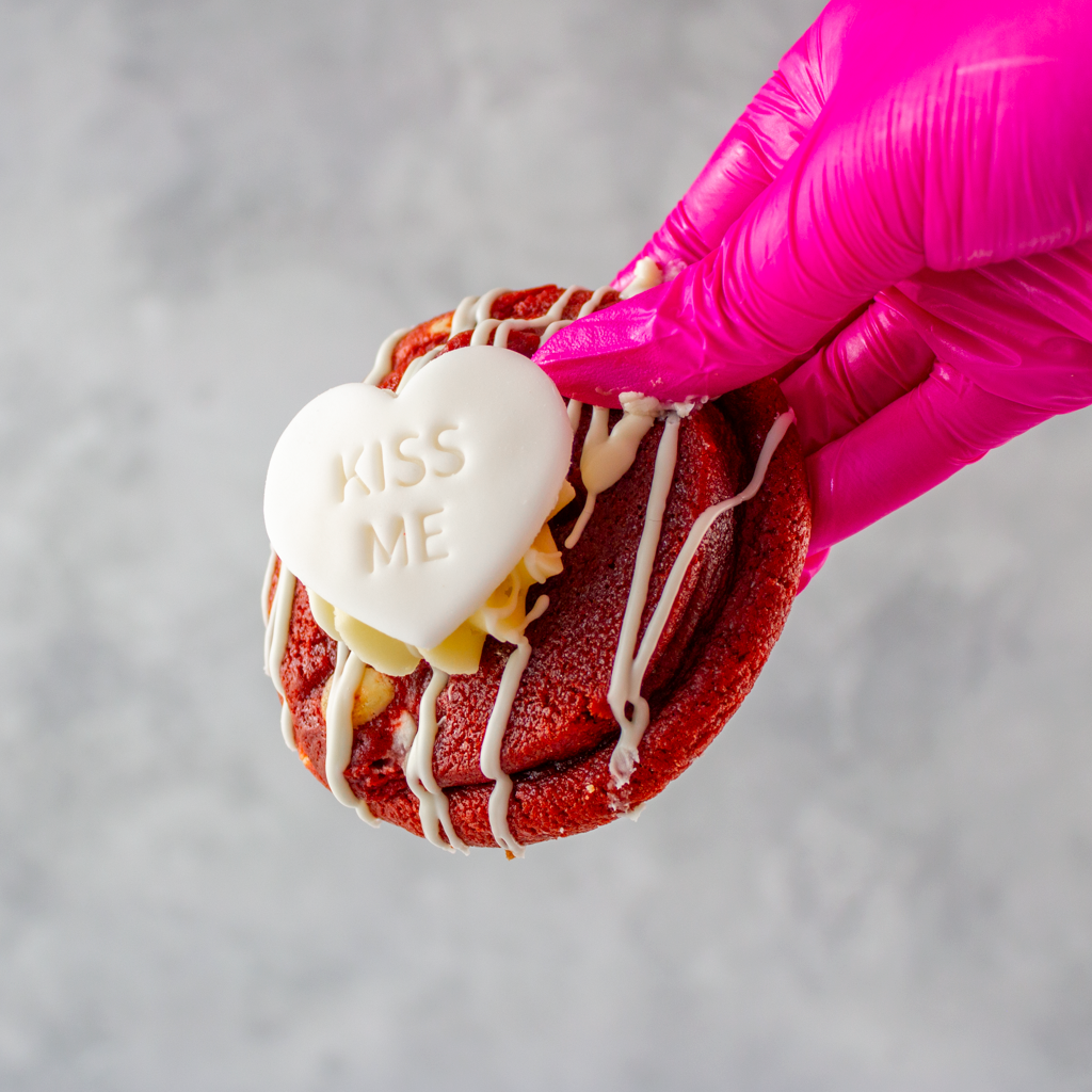 DoughGirl red velvet deluxe cookie with a heart-shaped 'Kiss Me' cookie, held by a pink gloved hand against a gray background.