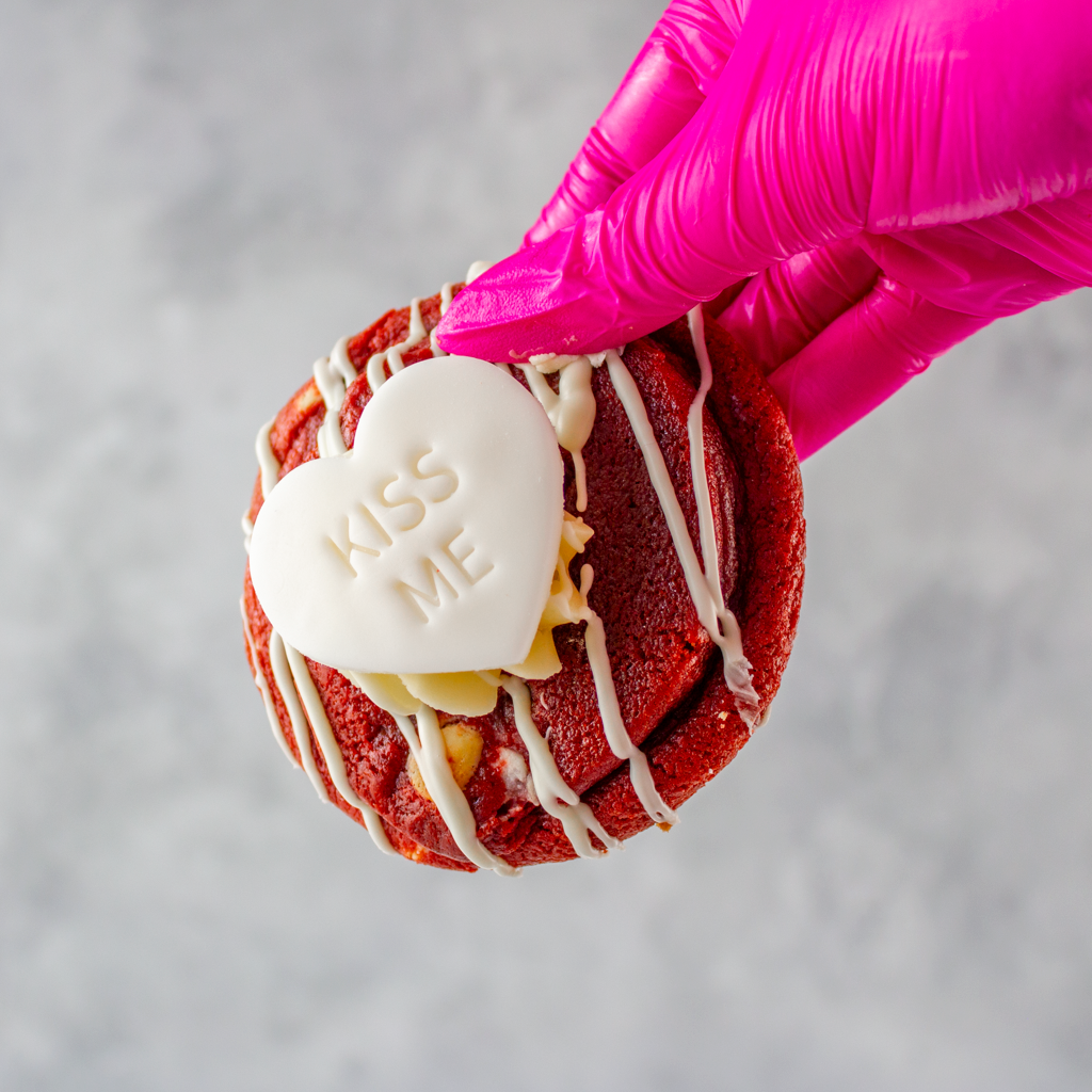 Limited Edition Valentine's Red Velvet Deluxe Cookie with white icing and a heart-shaped candy labeled 'Kiss Me' held by a pink gloved hand on a gray background.