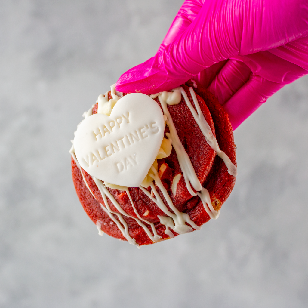 Limited Edition Valentine's Red Velvet Deluxe Cookie with white chocolate drizzle and a heart-shaped 'Happy Valentine's Day' candy, held by a pink gloved hand.