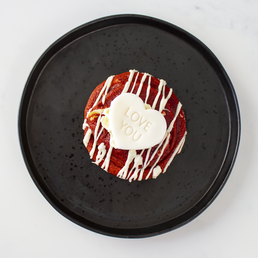 Red velvet cookie by DoughGirl with white drizzle and 'I LOVE YOU' heart on a black plate.