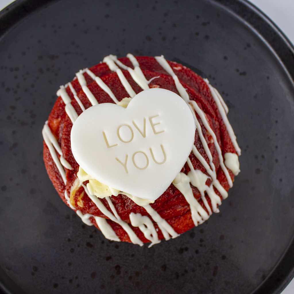 Red velvet deluxe cookie with white chocolate drizzle and a heart-shaped candy reading 'Love You' on a black plate.