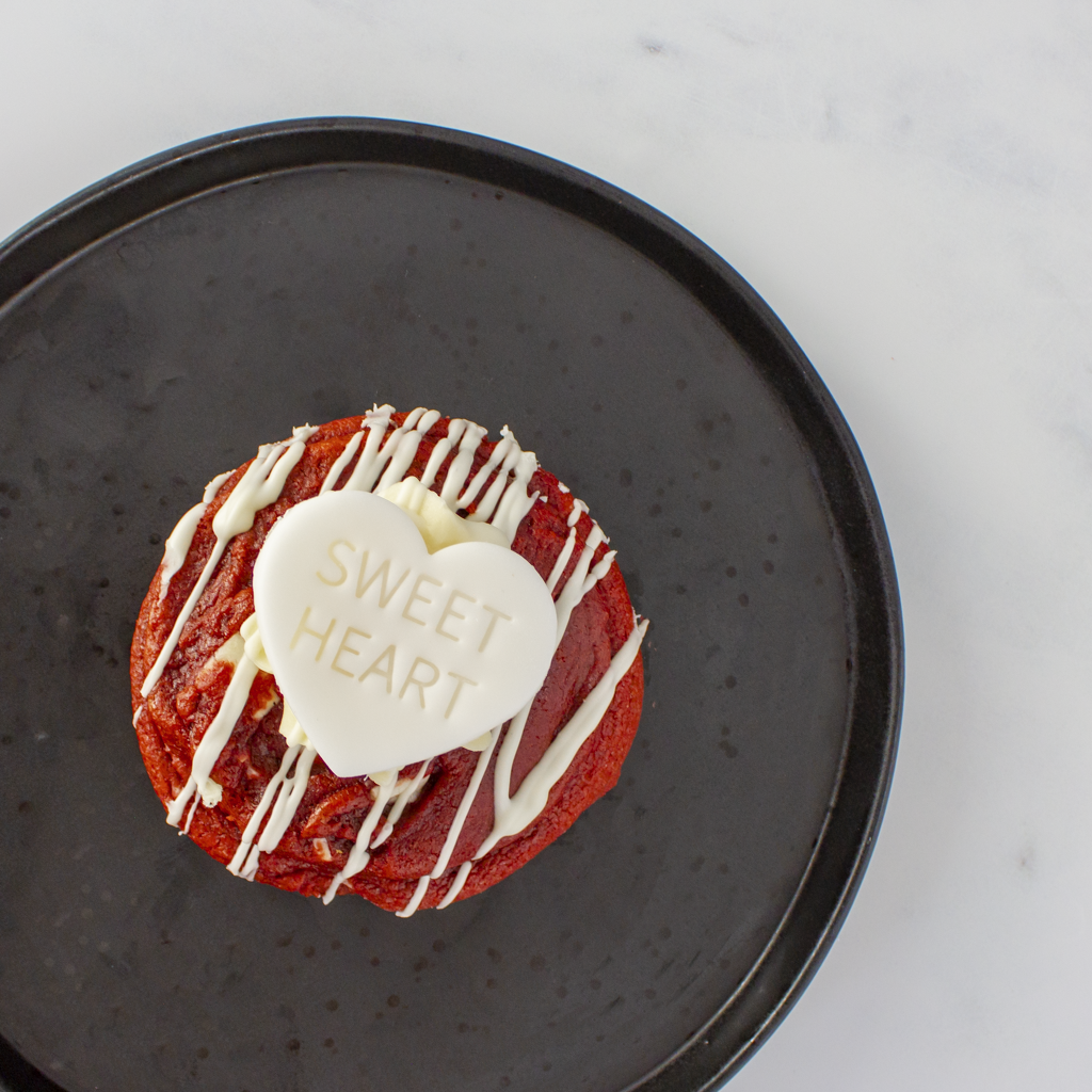 Red velvet DoughGirl cookie pie with white icing and a "sweet heart" heart-shaped white chocolate on a black plate.