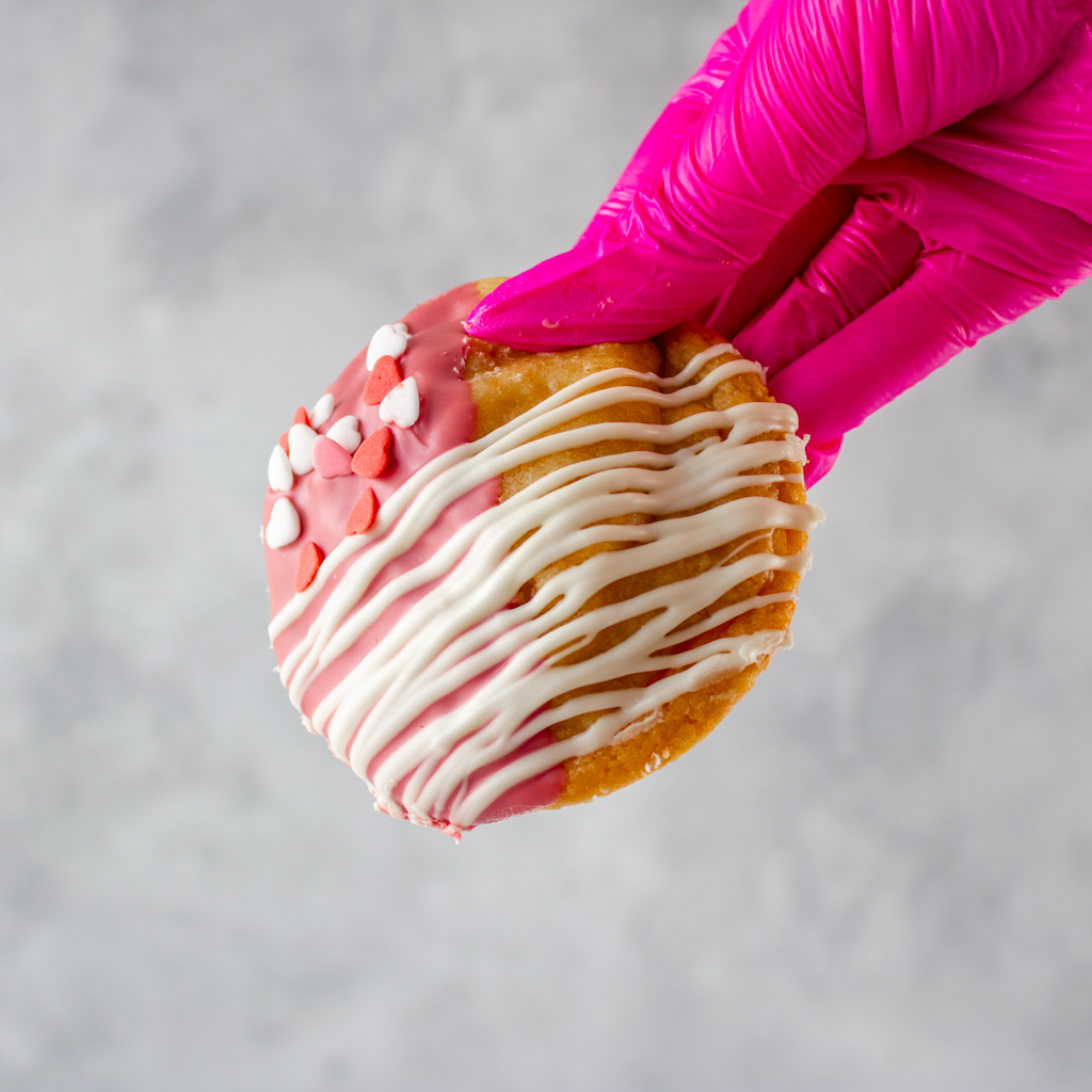 DoughGirl Valentine's Ruby Chocolate Deluxe Cookie with pink berry-like and white icing held by a pink gloved hand against a gray background