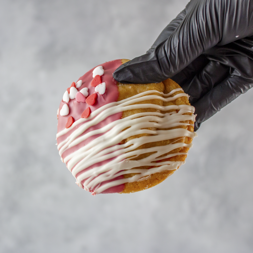 Heart-shaped cookie with pink and white icing held by a black gloved hand against a gray background