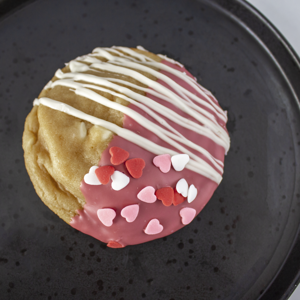 Cookie with pink and white heart sprinkles and white drizzle on a black plate