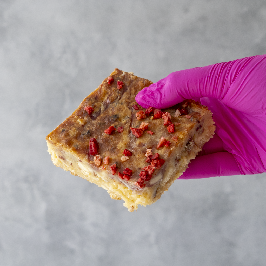 Hand holding a DoughGirl Eton Mess Blondie Slab with crunchy meringue pieces and sweet dried strawberry toppings against a gray background