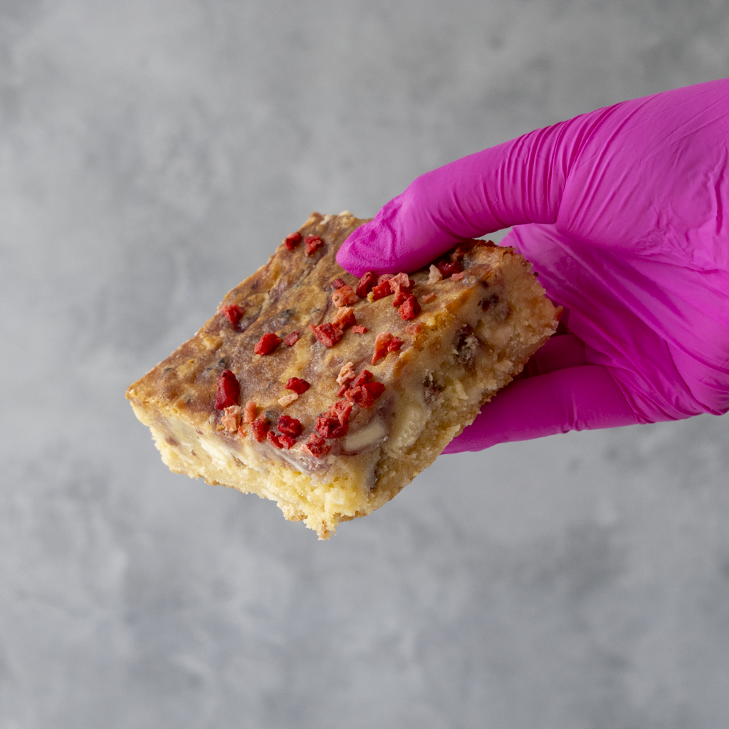 Hand wearing a pink glove holding a Eton Mess Blondie Slab by DoughGirl UK with fruity toppings against a gray background