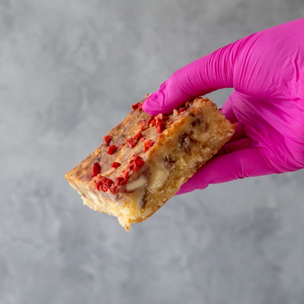 Hand wearing a pink glove holding a Eton Mess Blondie Slab with red toppings against a gray background