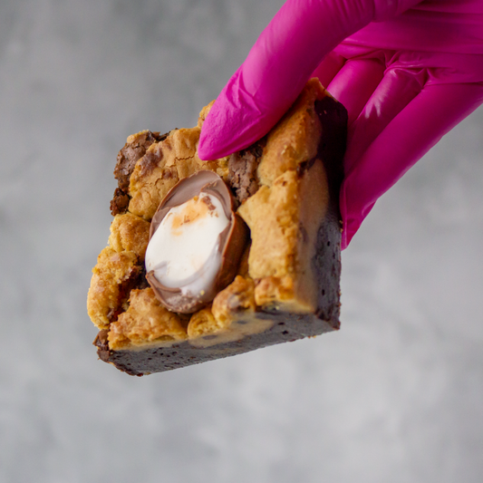 Hand wearing a pink glove holding a DoughGirl Creme Egg Brookie Slab with a visible filling against a gray background