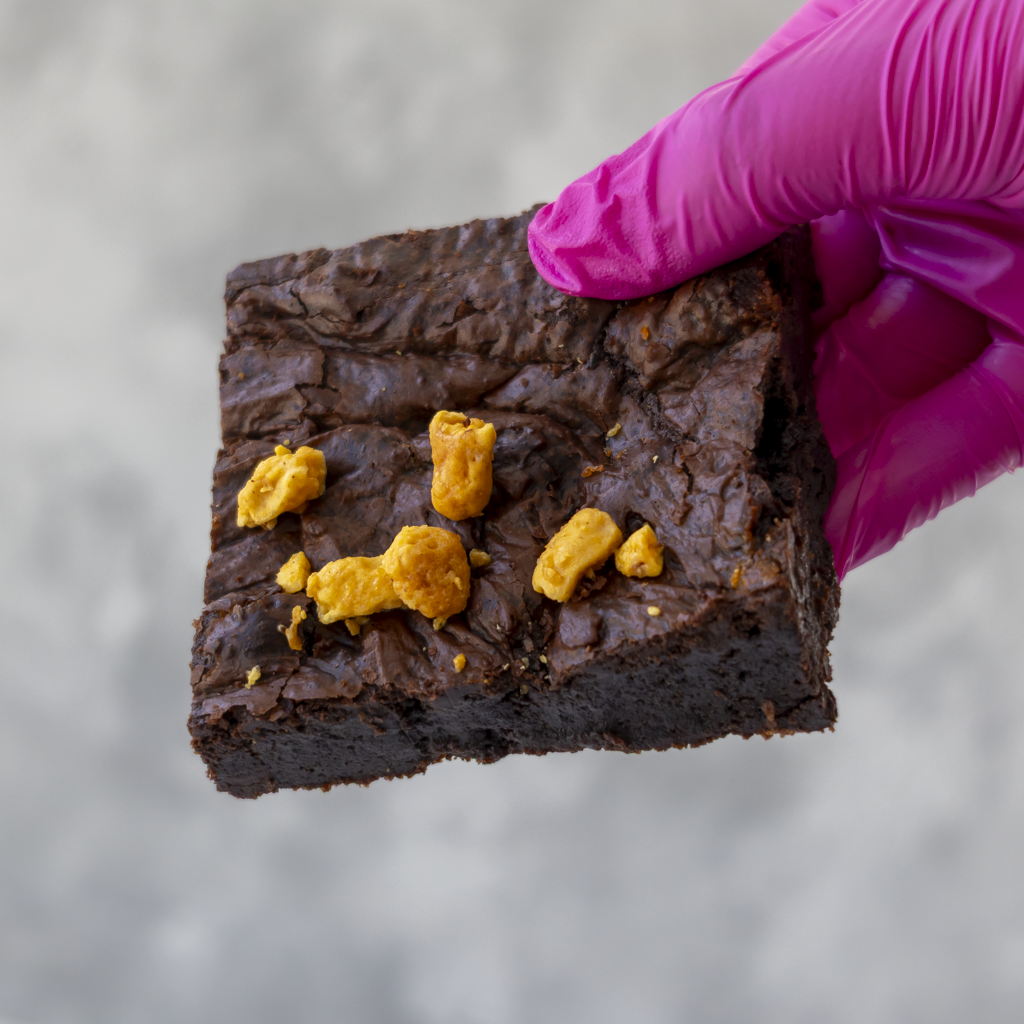A hand wearing a bright pink glove holds a square dark brown chocolate brownie topped with several crunchy honeycomb pieces, set against a soft gray background.