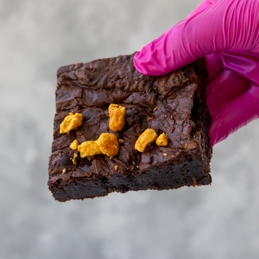 A hand wearing a bright pink glove holds a square dark brown chocolate brownie topped with several crunchy honeycomb pieces, set against a soft gray background.