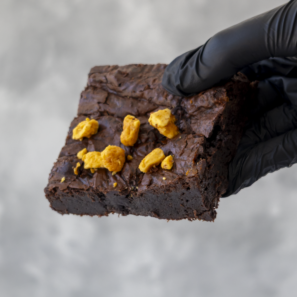 A hand wearing a black glove holds a square brownie topped with crunchy honeycomb against a soft gray background.