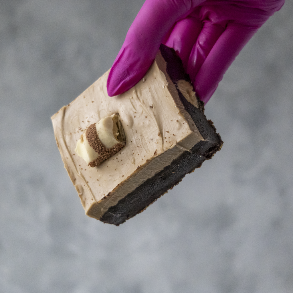 Bueno Brownie Slab by DoughGirl UK, with hazelnut chocolate spread and Kinder Bueno pieces, being held by a hand wearing a purple glove on a gray background