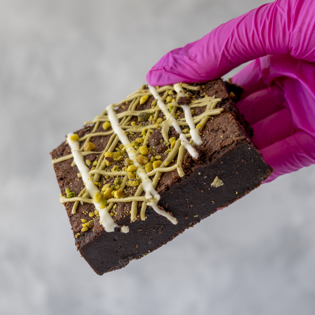 Pistachio Brownie Slab held in pink glove with white chocolate drizzle and pistachio topping
