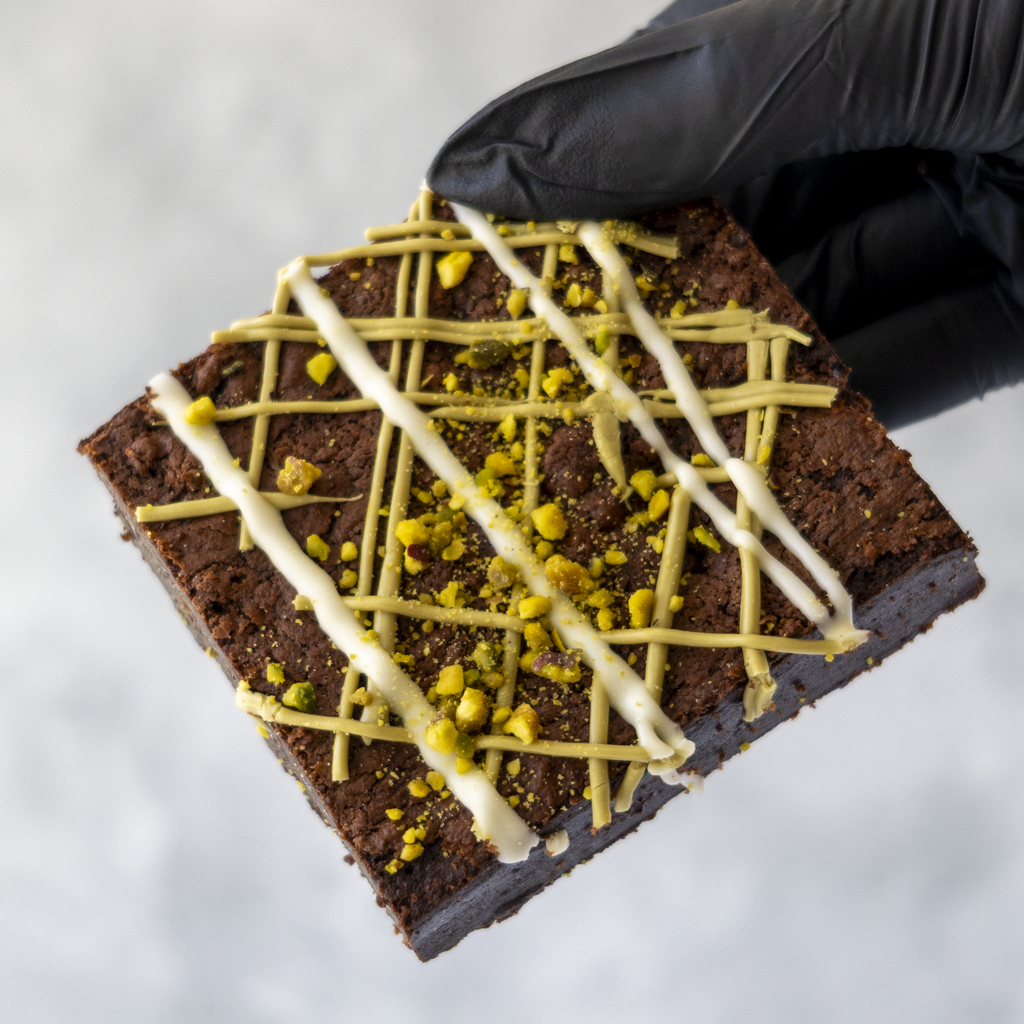 Pistachio Brownie Slab held in black glove with white chocolate drizzle and pistachio topping