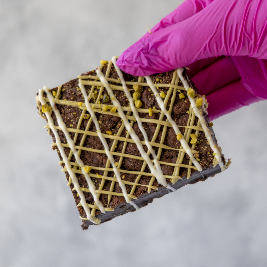 Pistachio Brownie Slab held in pink glove with crushed pistachios and drizzle topping