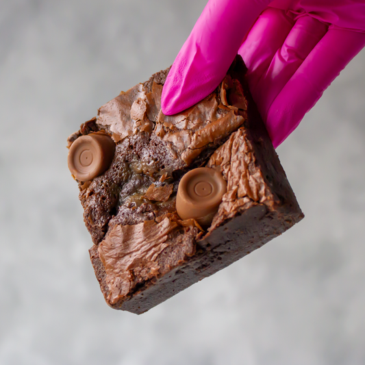 DoughGirl Rolo Brownie Slab with Rolo milk chocolate & caramel, held by a hand wearing a pink glove on a gray background