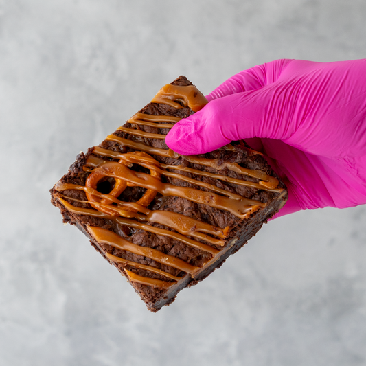 Salted Caramel Pretzel Brownie Slab with caramel drizzle held by a hand wearing a pink glove on a gray background